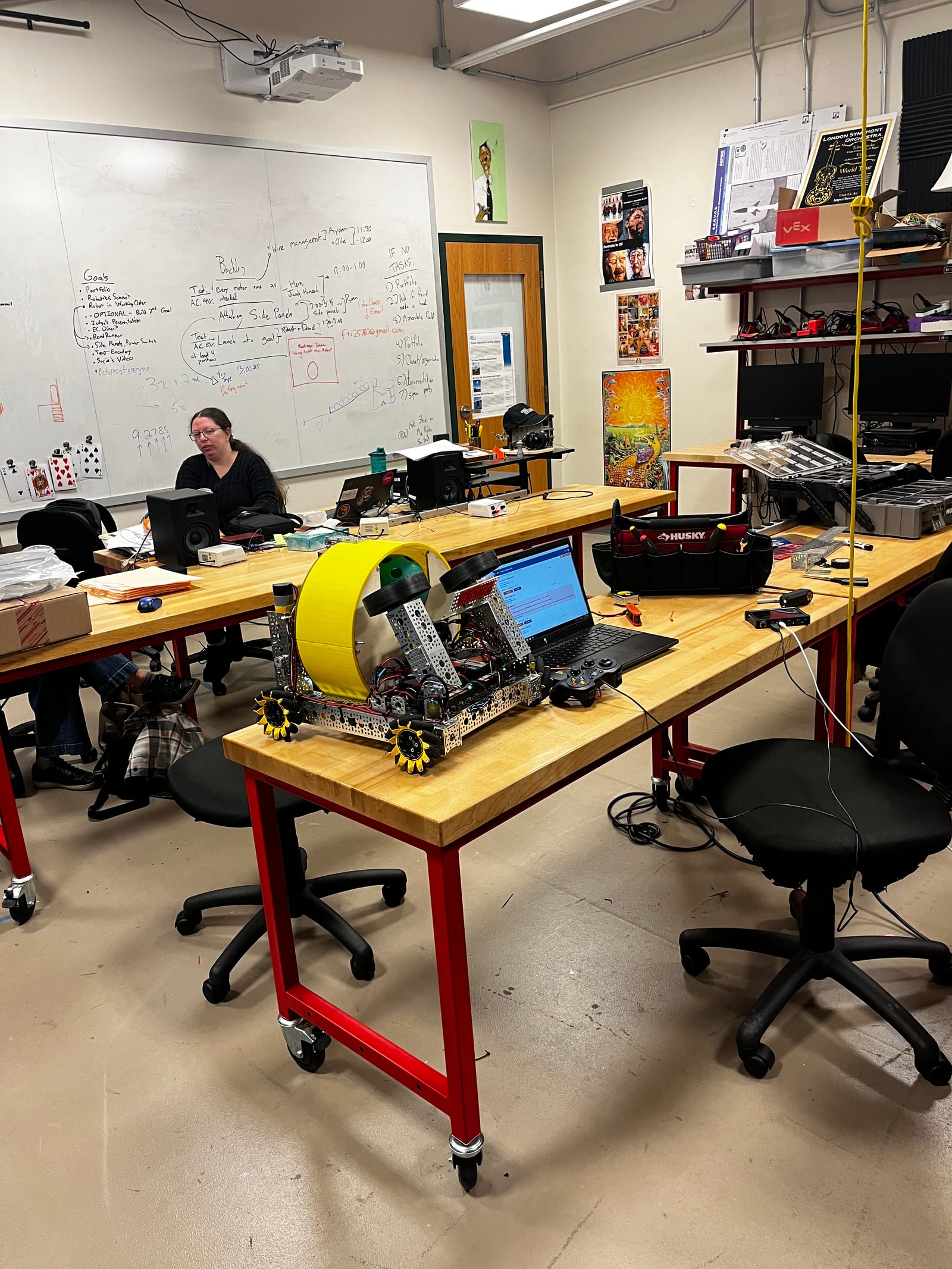 The robot on a workbench in the shop with the day's goals on the whiteboard